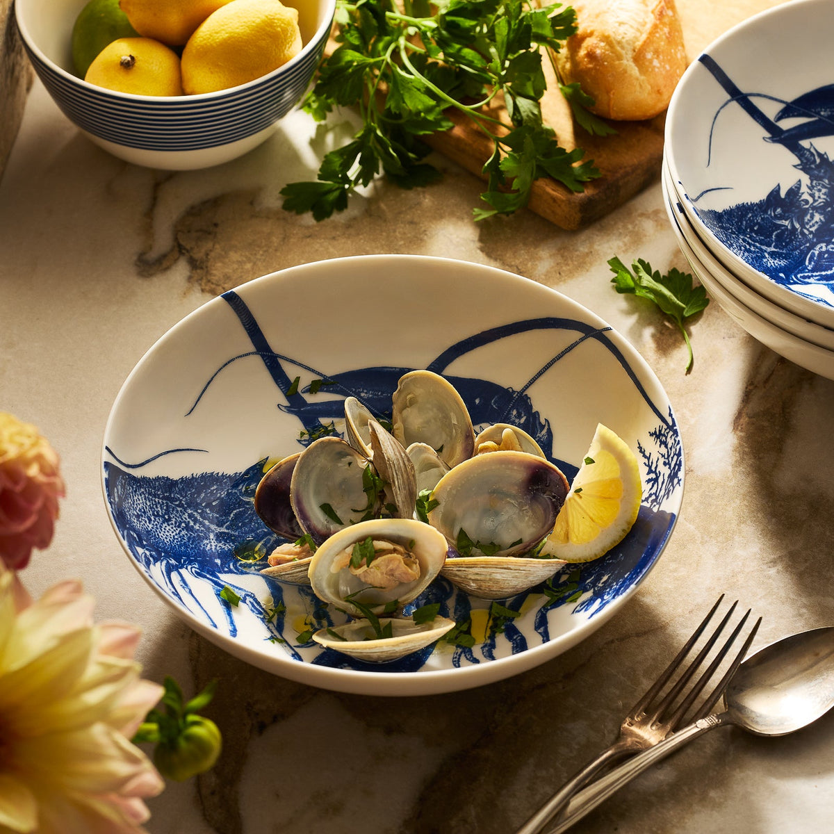 The Caskata Lobster Entrée Bowl, shown with clams, parsley, and lemon on marble, comes in elegant porcelain with a fork, flowers, and fresh herbs nearby. This beautiful bowl is also dishwasher safe for easy cleanup.