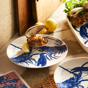A baked stuffed clam with breadcrumbs sits on a Caskata Lobster Coupe Salad Plate, topped with a lemon wedge. Two whole lemons and fresh greens in the background bring vibrant color to the scene.