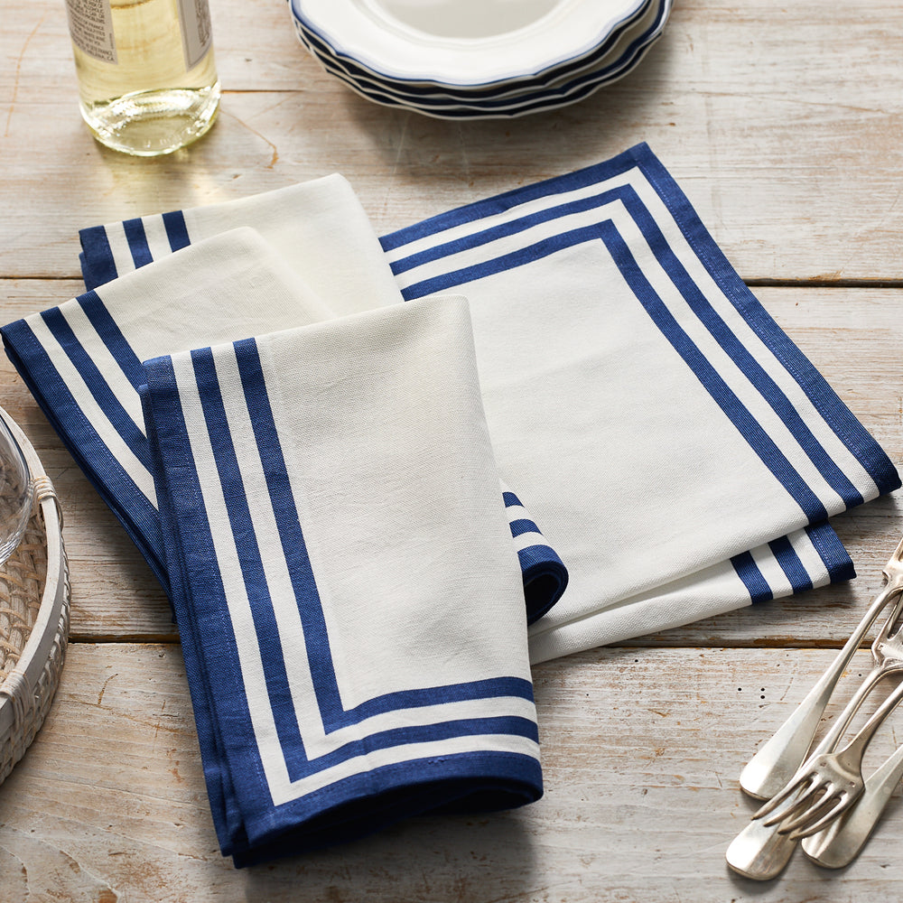 Two Caskata Triple Stripe Blue Dinner Napkins are folded on a wooden table beside stacked plates, cutlery, and a bottle of white wine.