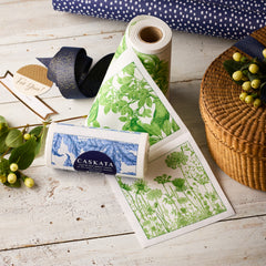 MY DRAP Mixed Botanical Cocktail Napkin Rolls, Set of 2, are shown on a rustic white table with a woven basket, ribbon, and gift tag—stylish and eco-friendly for gatherings.
