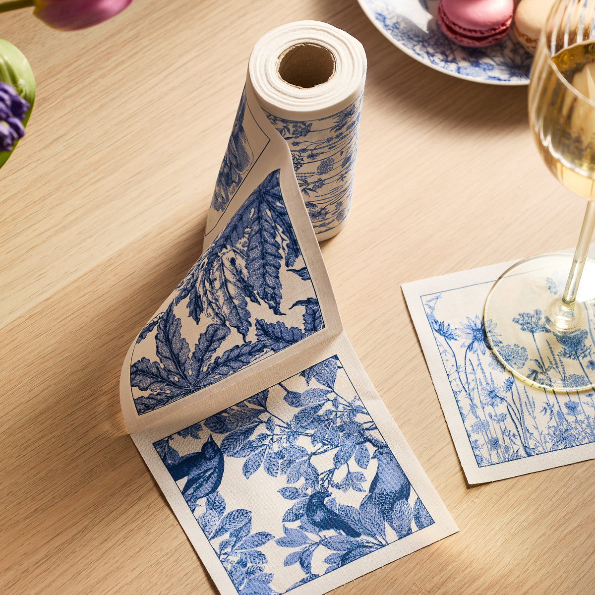 The "Arbor Coffee Talk" Bundle by Caskata features blue and white botanical cocktail napkins, partially unrolled on a light wooden table beside a plate of macarons and a glass of white wine.