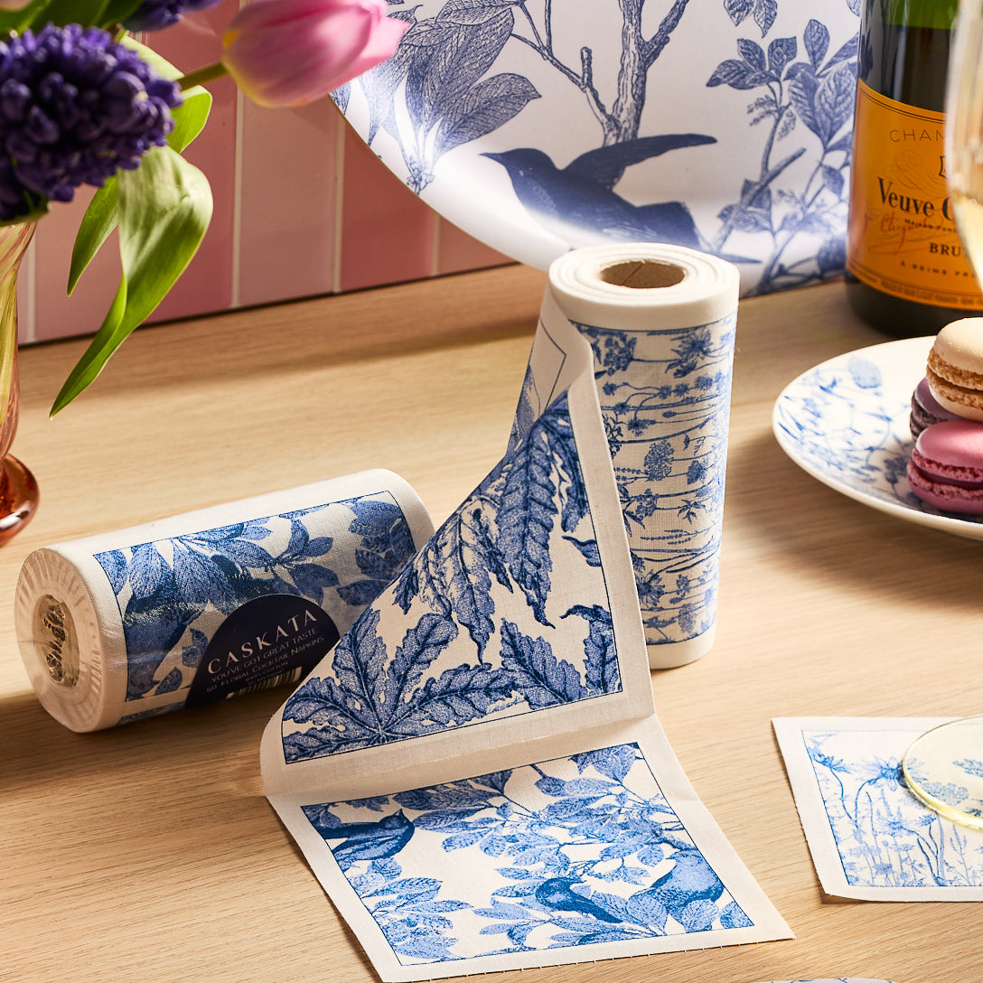 A roll of MY DRAP’s Botanicals Cocktail Napkin Roll is unspooled, with three individual square napkins featuring blue and white floral and botanical designs placed beside it on a beige surface—evoking the charming motifs often found on handcrafted cotton napkins.