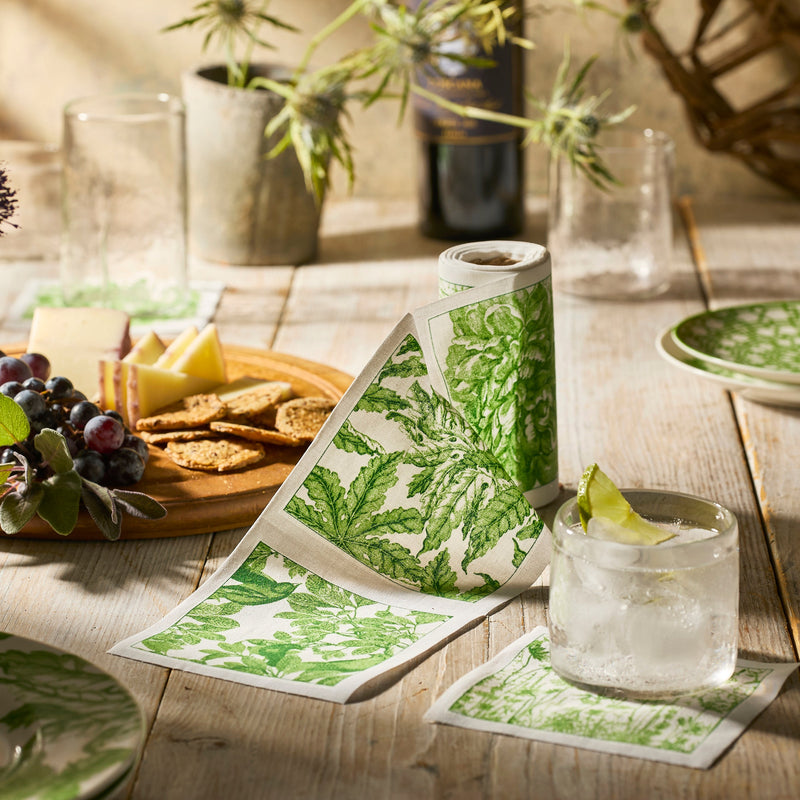 A glass of ice water with lime rests on a leafy-patterned napkin beside the MY DRAP Botanicals Verde Cocktail Napkin Roll and a cheese board with crackers, grapes, and cheese, all arranged on a wooden table.