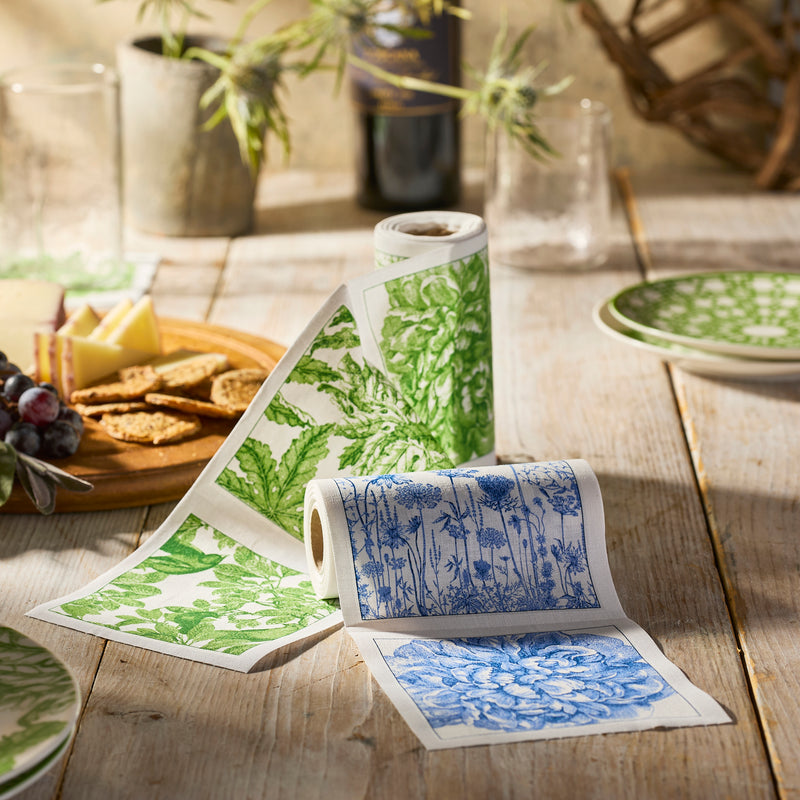 Two MY DRAP Botanicals Verde Cocktail Napkin Rolls, featuring green and blue botanical prints, are unrolled on a wooden table set with plates, glasses, crackers, cheese, and grapes.