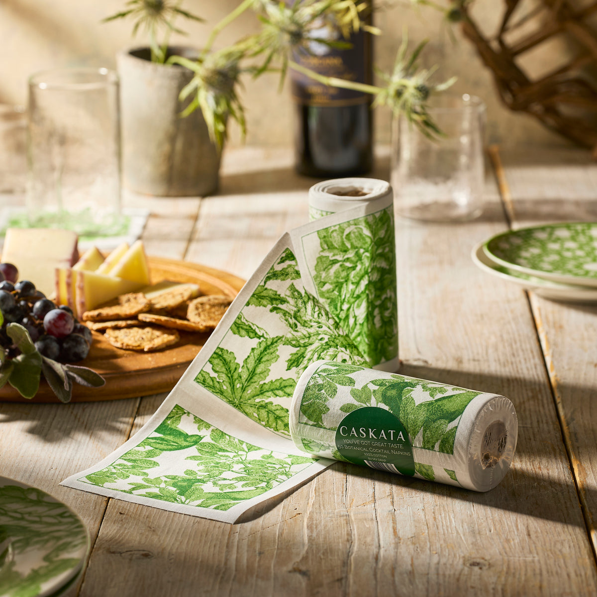 The "Freya Coffee Talk" Bundle by Caskata features two green and white botanical napkin rolls on a rustic table with cheese, grapes, wine, and glassware—an elegant nature-inspired gift for any occasion.