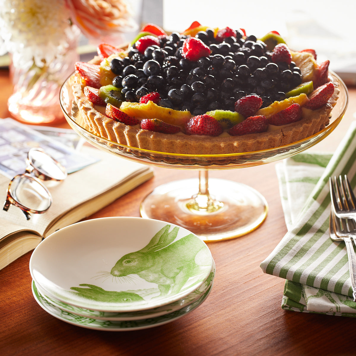 A fruit tart on a glass cake stand, surrounded by a stack of Caskata Bunnies Verde Small Plates, a folded green-striped napkin, and a pair of eyeglasses on an open book.