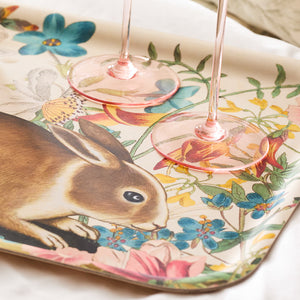Two glasses with pink stems sit elegantly on the Caskata Bunny Maximus Large Rectangular Birchwood Tray, featuring a whimsical rabbit and vibrant flowers.