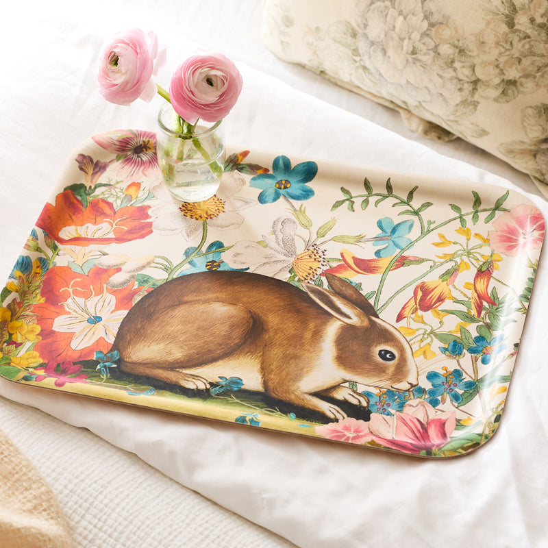 The Bunny Maximus Large Rectangular Birchwood Tray by Caskata, crafted sustainably, showcases a charming rabbit and flower design. The tray holds two pink flowers in a glass vase and is set against a bed with floral pillows.