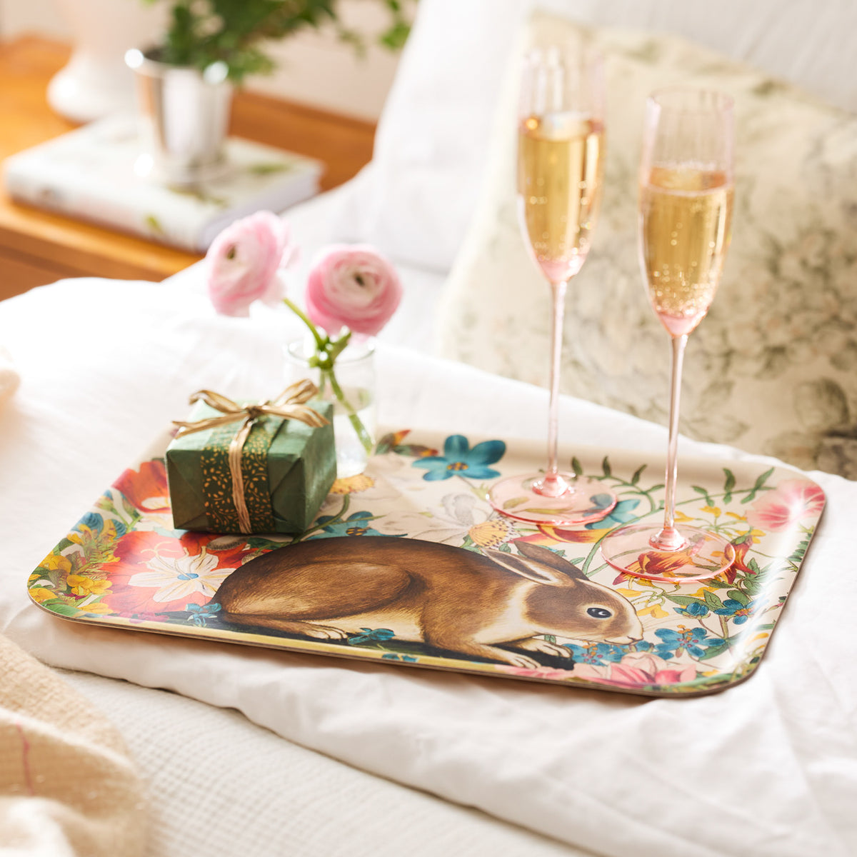 The Bunny Maximus Large Rectangular Birchwood Tray by Caskata features a rabbit illustration and holds two champagne glasses, a vase of pink flowers, and a wrapped gift box on a bed.