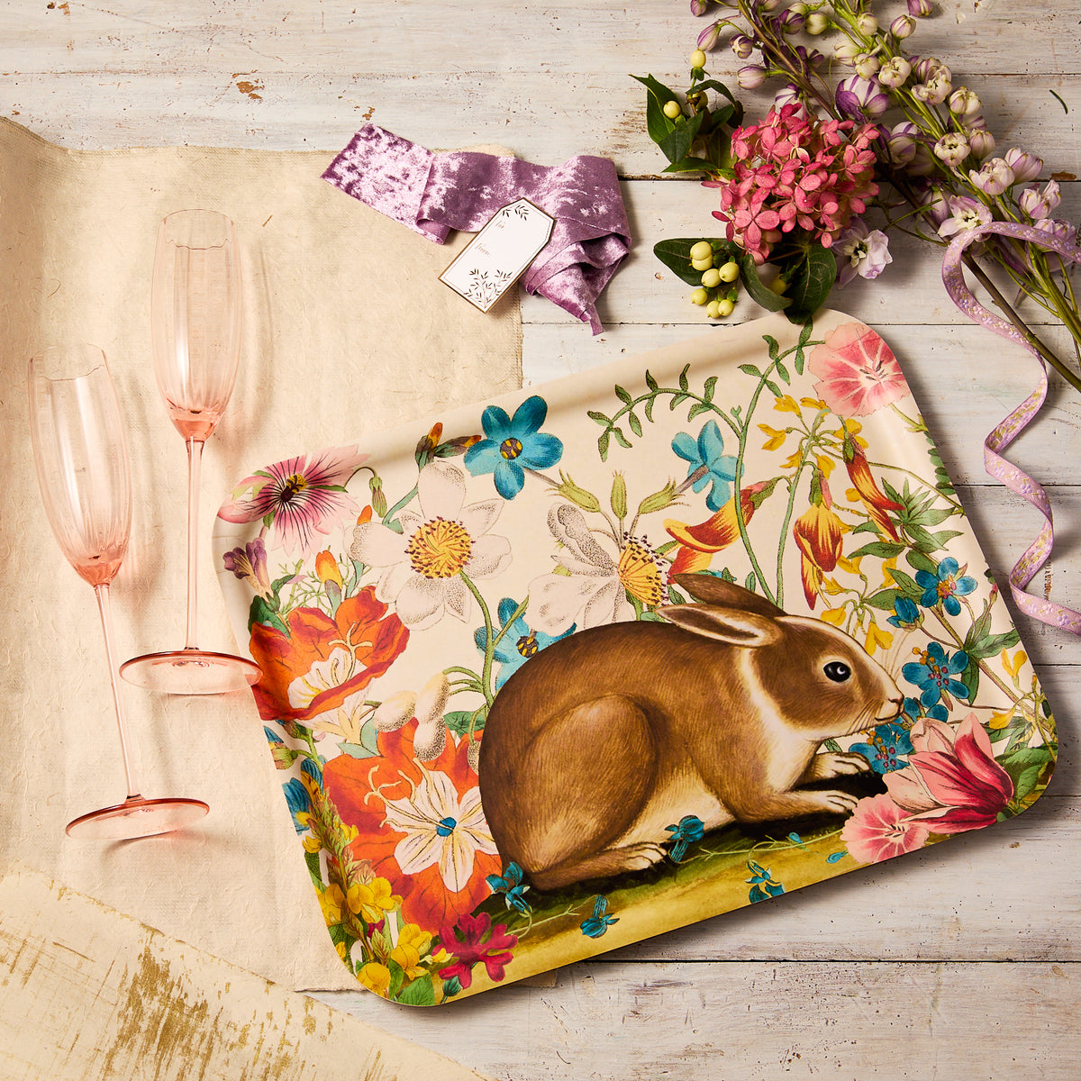 The "Breakfast in Bed" Bundle by Caskata features a handcrafted birchwood tray with a whimsical rabbit illustration, displayed alongside two pink glasses, a purple ribbon, and flowers on a light wood surface.