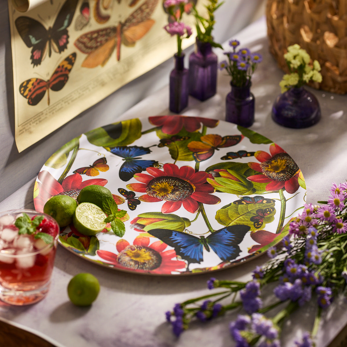 The Butterflies Large Round Birchwood Tray by Caskata, featuring a vibrant butterfly and floral design, is displayed on a table with limes, a red iced drink, small vases of flowers, and a butterfly poster in the background.