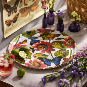 The Butterflies Large Round Birchwood Tray by Caskata, featuring a vibrant butterfly and floral design, is displayed on a table with limes, a red iced drink, small vases of flowers, and a butterfly poster in the background.