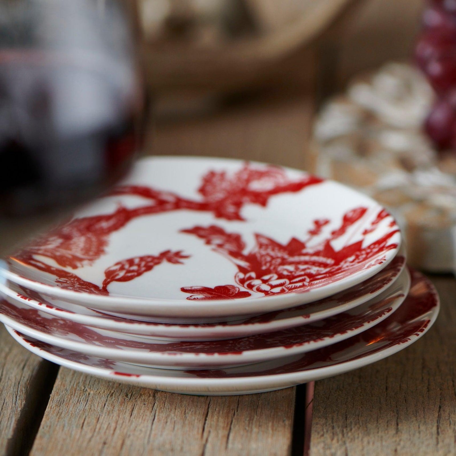 Three small crimson floral plates from Caskata’s Happy Hosting Bundle are stacked on wood, with blurred objects and a glass in the background—a charming accent for any gathering.