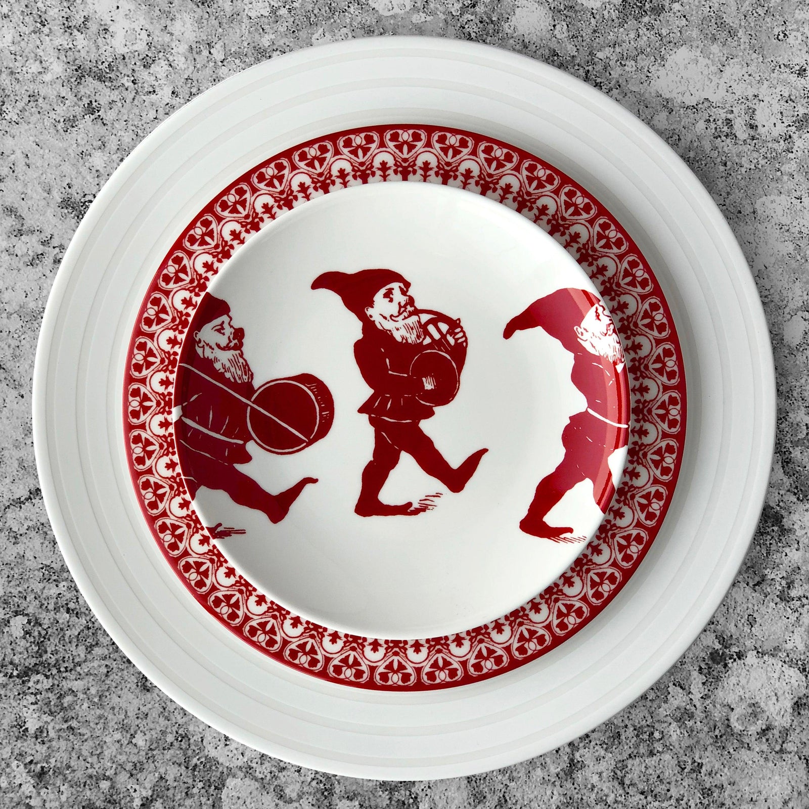 Four white plates, part of an heirloom-quality dinnerware holiday collection, each feature an illustration of a red gnome holding a drum. They are called Elves Small Plates by Caskata Artisanal Home.