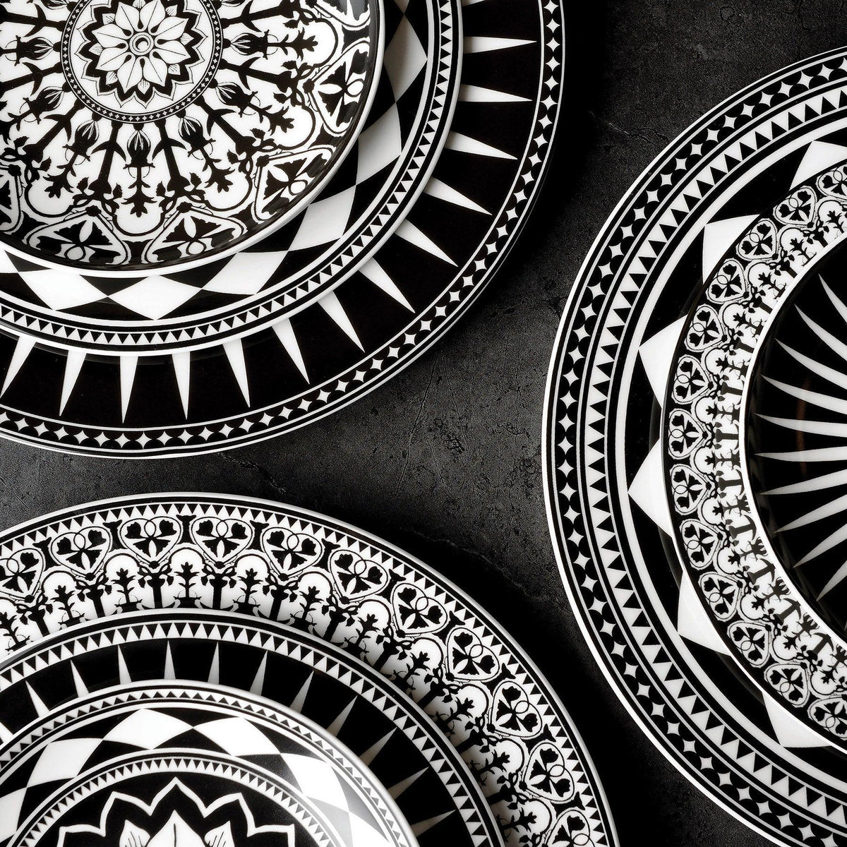 Four intricately patterned black and white Marrakech Rimmed Dinner Plates with geometric and graphic floral designs from Caskata Artisanal Home are placed on a dark surface.