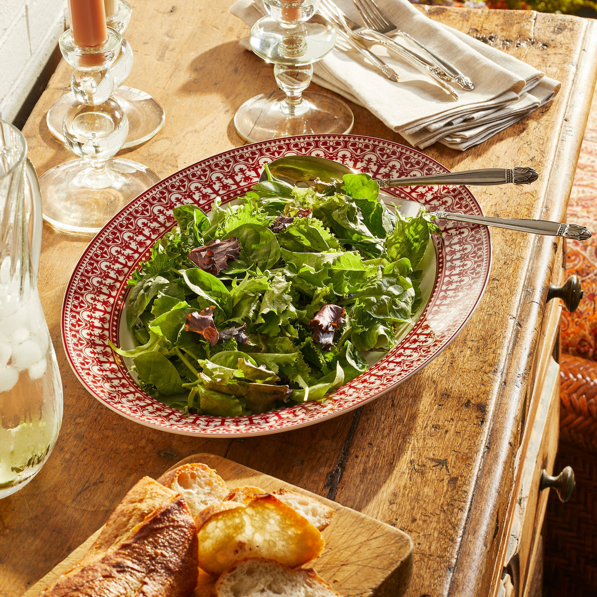 A bowl of mixed green salad on a stylish Casablanca Crimson Oval Rimmed Platter from Caskata Artisanal Home rests on a wooden table with a fork and spoon. Nearby are a glass of water with ice, a stack of folded napkins, candles, and a wooden board with sliced bread, creating an inviting Casablanca-inspired dining scene.