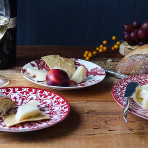 A table set with heirloom-quality Casablanca Crimson Small Plates by Caskata Artisanal Home features red patterned plates holding slices of cheese, bread, and a plum. In the background, there is a glass of white wine, a wine bottle, and a bunch of red grapes. A cheese knife completes this elegant arrangement.