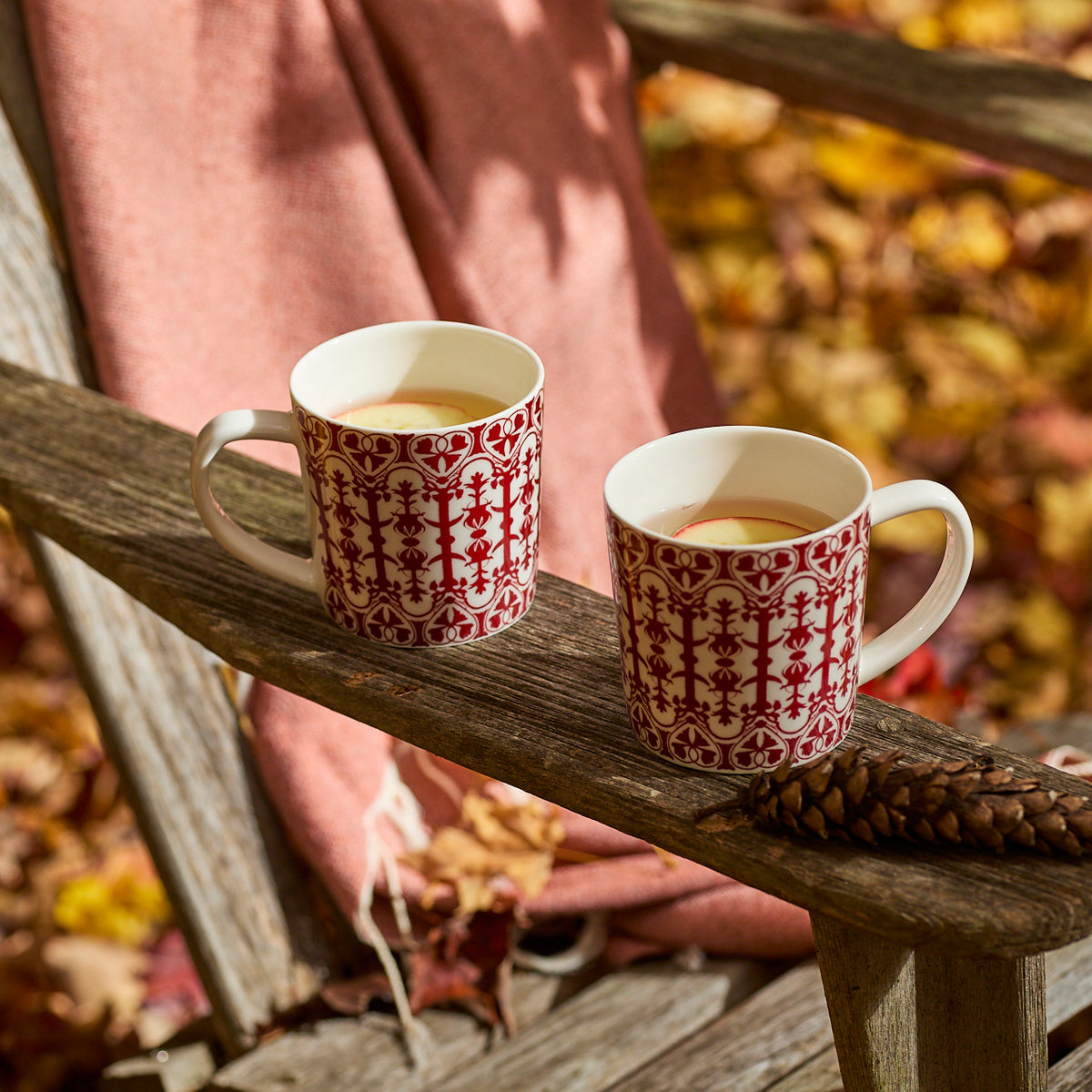 Two "Holiday Cocoa" Bundle porcelain mugs by Caskata filled with a light beverage sit on a wooden chair arm outdoors, surrounded by autumn leaves and a pine cone.