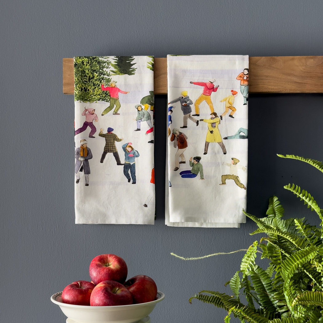 An illustration of people engaging in a snowball fight amidst winter scenery, dressed warmly, with snow-covered trees creating a backdrop reminiscent of the design on the Caskata X Felix Doolittle Snowball Fun Kitchen Towels, Set of 2.