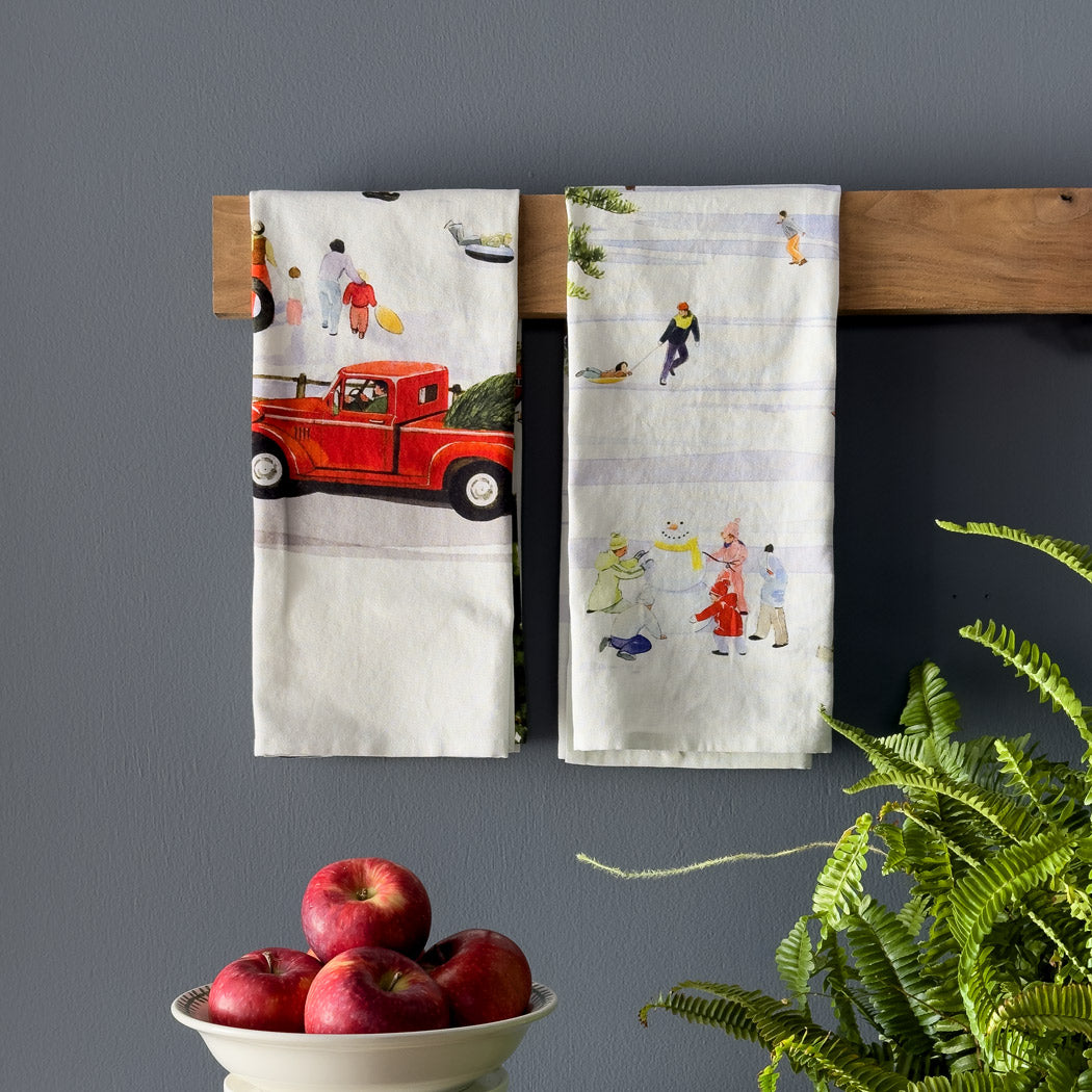 The Caskata X Felix Doolittle Winter Wonderland Kitchen Towels, Set of 2, showcase winter scenes such as skating, sledding, building a snowman, and a red truck transporting holiday cargo.