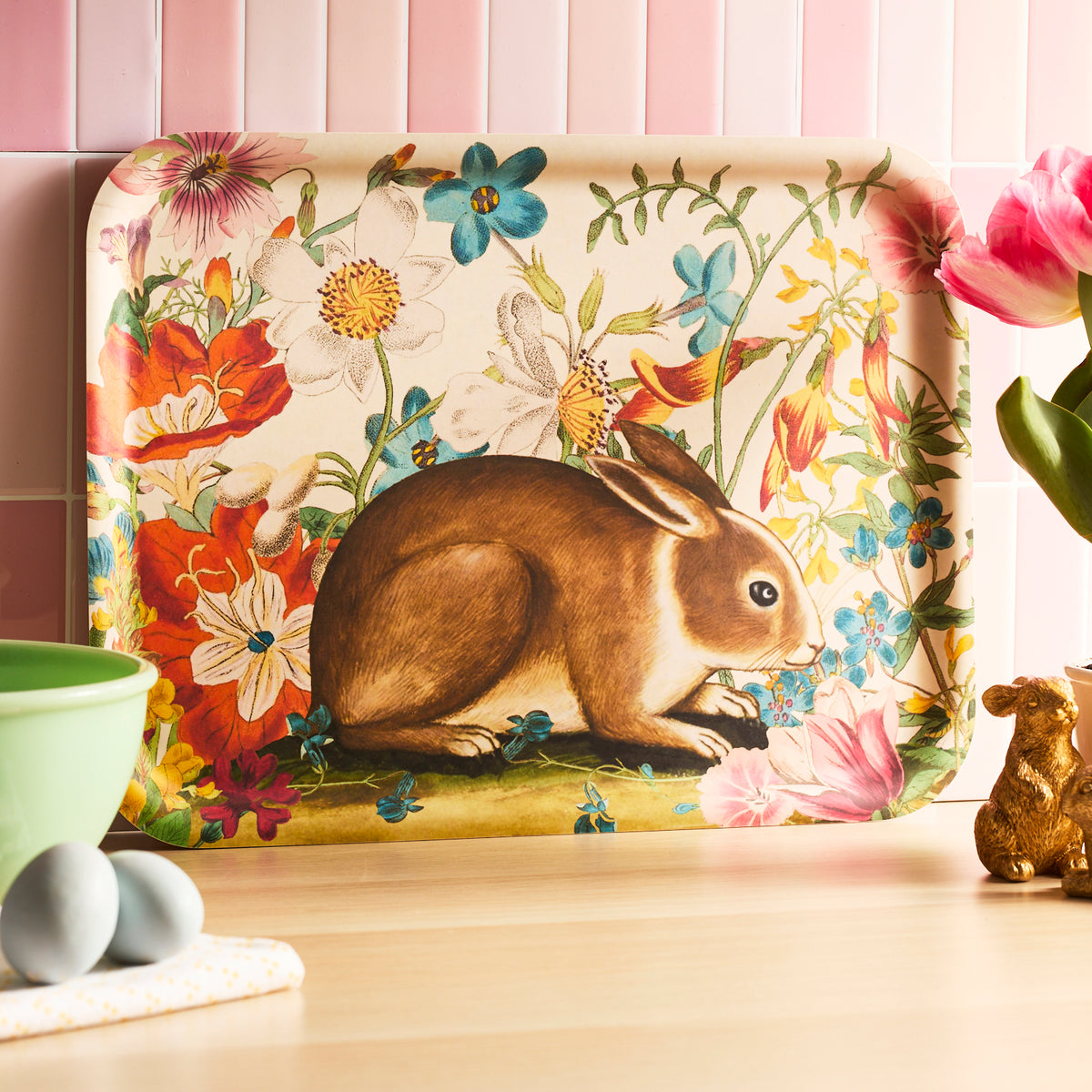 The Bunny Maximus Large Rectangular Birchwood Tray by Caskata showcases a brown rabbit amid colorful flowers on a light wood surface, crafted from sustainable birchwood. It&#39;s perfect for adding charm to any kitchen setting with pink tiles in the background.