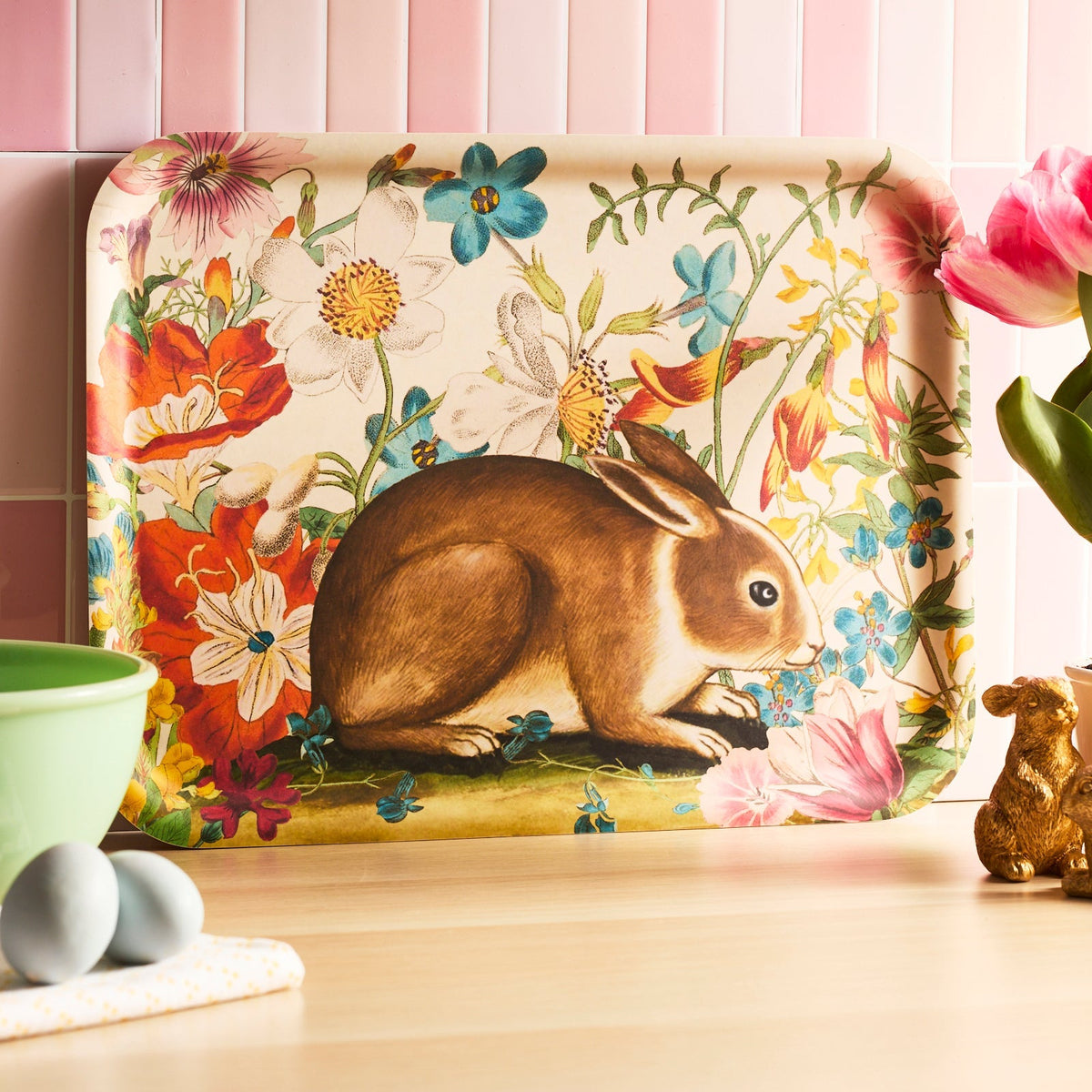 The Bunny Maximus Large Rectangular Birchwood Tray by Caskata showcases a brown rabbit amid colorful flowers on a light wood surface, crafted from sustainable birchwood. It's perfect for adding charm to any kitchen setting with pink tiles in the background.