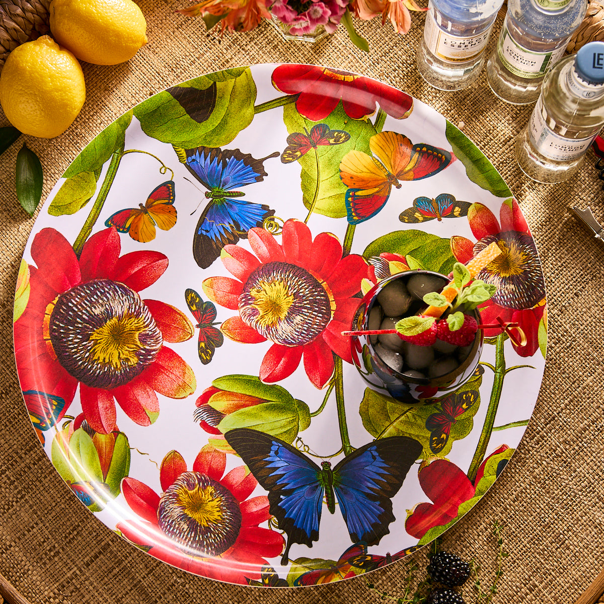 The Caskata Butterflies Large Round Birchwood Tray, featuring red flowers and blue butterflies, holds a cocktail with fruit and herbs. Lemons and bottles rest on a woven surface, adding natural charm to this sustainable setting.