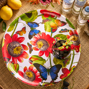 The Caskata Butterflies Large Round Birchwood Tray, featuring red flowers and blue butterflies, holds a cocktail with fruit and herbs. Lemons and bottles rest on a woven surface, adding natural charm to this sustainable setting.