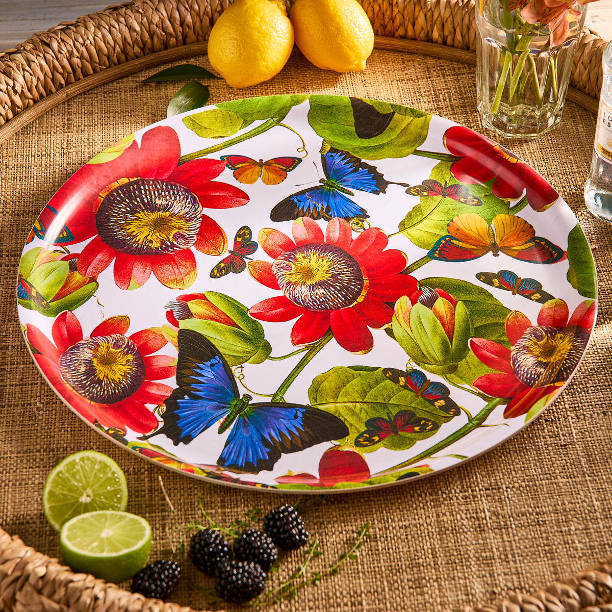 The Butterflies Large Round Birchwood Tray by Caskata features a vibrant design of red flowers, green leaves, and colorful butterflies on a woven surface with lemons, a glass, lime slices, and blackberries, adding an artistic flourish to sustainable dining moments.