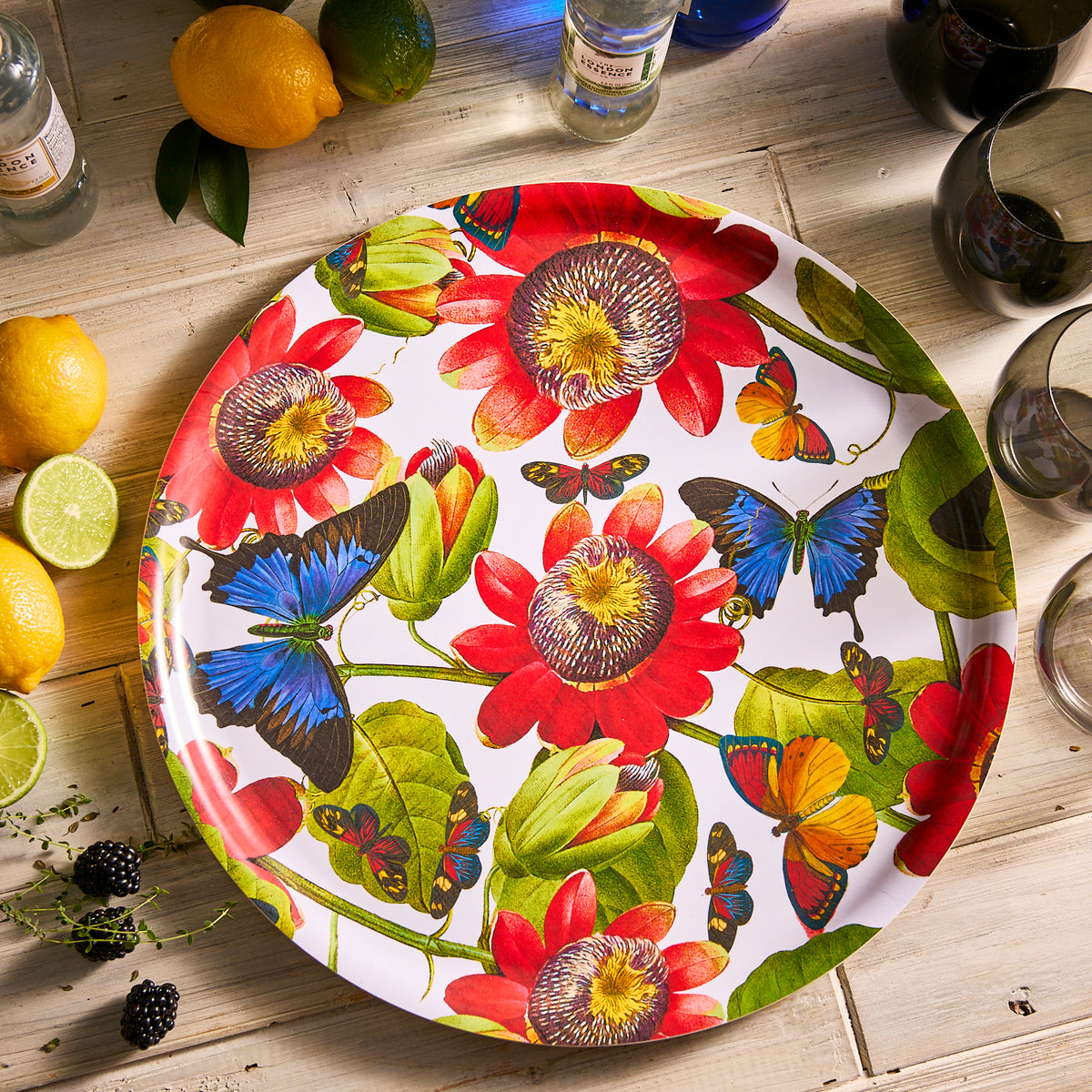 The vibrant Caskata Butterflies Large Round Birchwood Tray, featuring red flowers, green leaves, and blue and orange butterflies, rests on a wooden table surrounded by lemons, limes, and glasses.