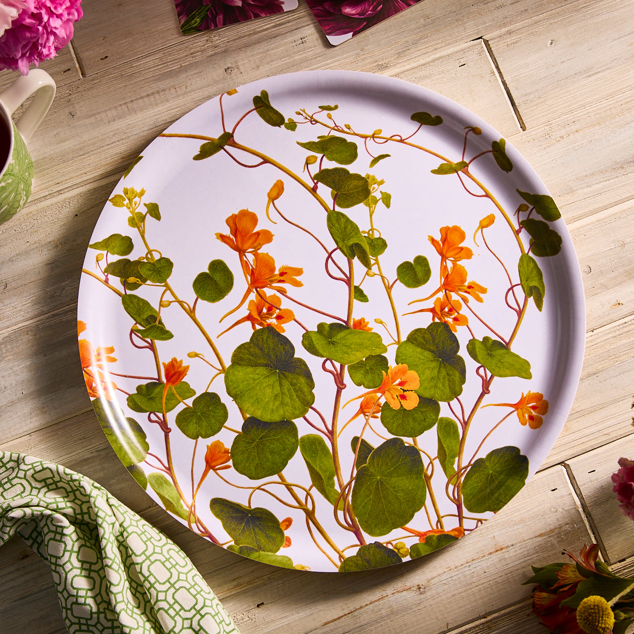 The Caskata "Serving Spring" Bundle features a round plate with an orange flower and green leaf pattern behind four porcelain mugs with green-and-white designs—a charming gift for gardeners.