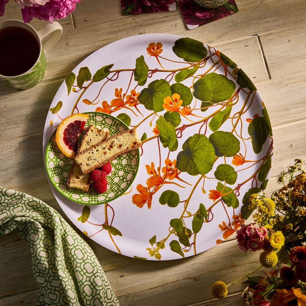 The Caskata Nasturtiums Large Round Birchwood Tray, adorned with an orange floral design, holds a green patterned plate filled with sliced bread and raspberries. Nearby, a cup of tea and flowers complete the eco-friendly ambiance on the wooden table.