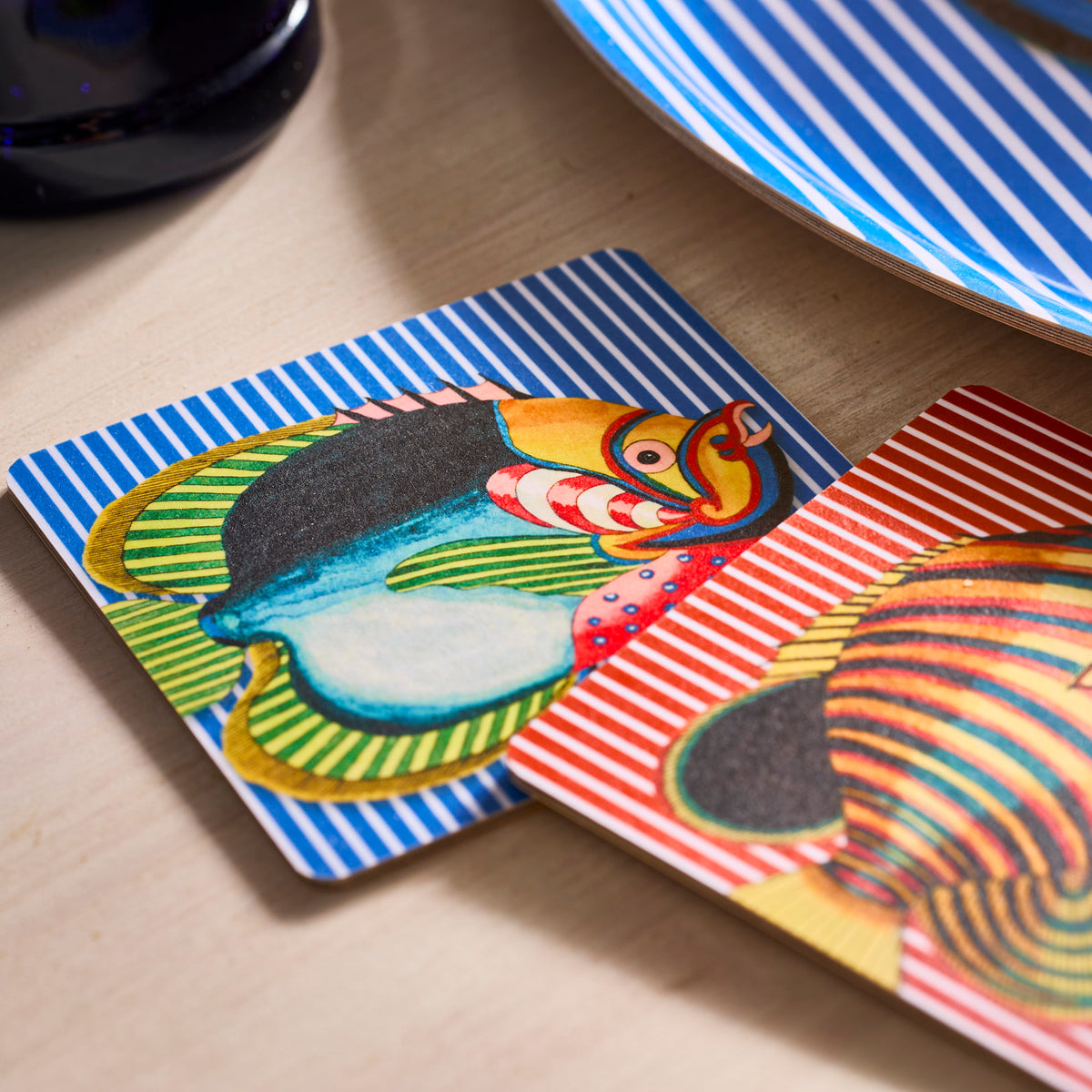 Two vibrant coasters from the Caskata Party Fish Birchwood Coasters Set rest on wood, looking like fish near a striped blue and white plate.