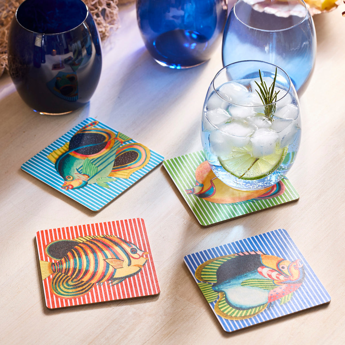 The Party Fish Birchwood Coasters by Caskata, a set of four vibrant and eco-friendly trays, lay on the table like a party under the sea. A cooling iced drink sits on one coaster, while nearby three blue glasses await the fun. Perfect for adding charm and eco-consciousness to any gathering!.