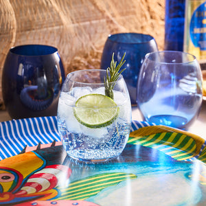 A glass of sparkling water with ice, lime, and rosemary sits on a colorful tray as the Les Nuages Blue Ombré Glasses Set of 4 by Caskata gleam elegantly in the background.
