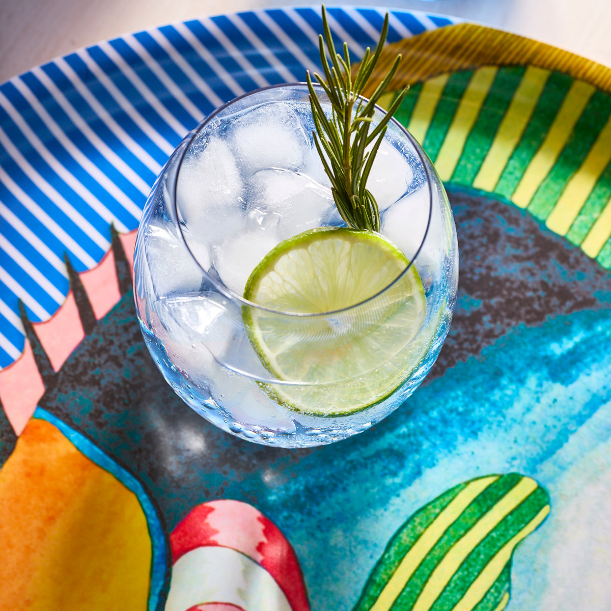 A Les Nuages Blue Ombré Glass from Caskata, filled with ice, a lime slice, and garnished with rosemary, sits on a colorful tray.