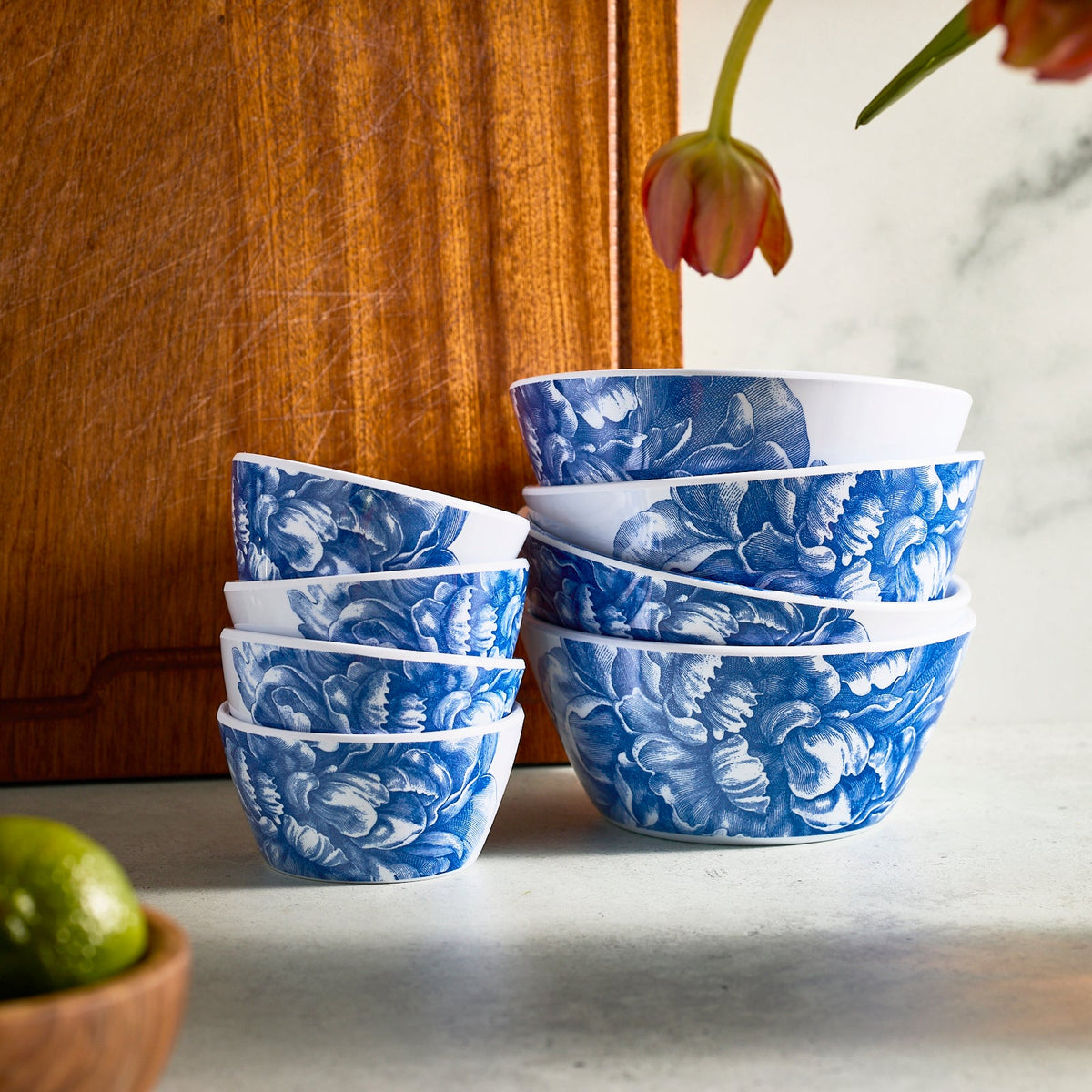On a kitchen counter, a set of Caskata Peony Melamine Cereal Bowls showcases elegance with blue and white floral patterns. BPA-free and ideal for indoor or outdoor use, their varying sizes complement a wooden board and limes nearby, blending practicality with style.