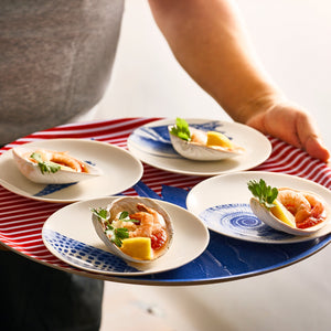 A person carries the Horseshoe Crab Large Round Birchwood Tray by Caskata, adorned with four plates showcasing seafood dishes garnished with lemon and parsley. Each plate features red and blue patterns, reminiscent of Finland's rich Nordic design.