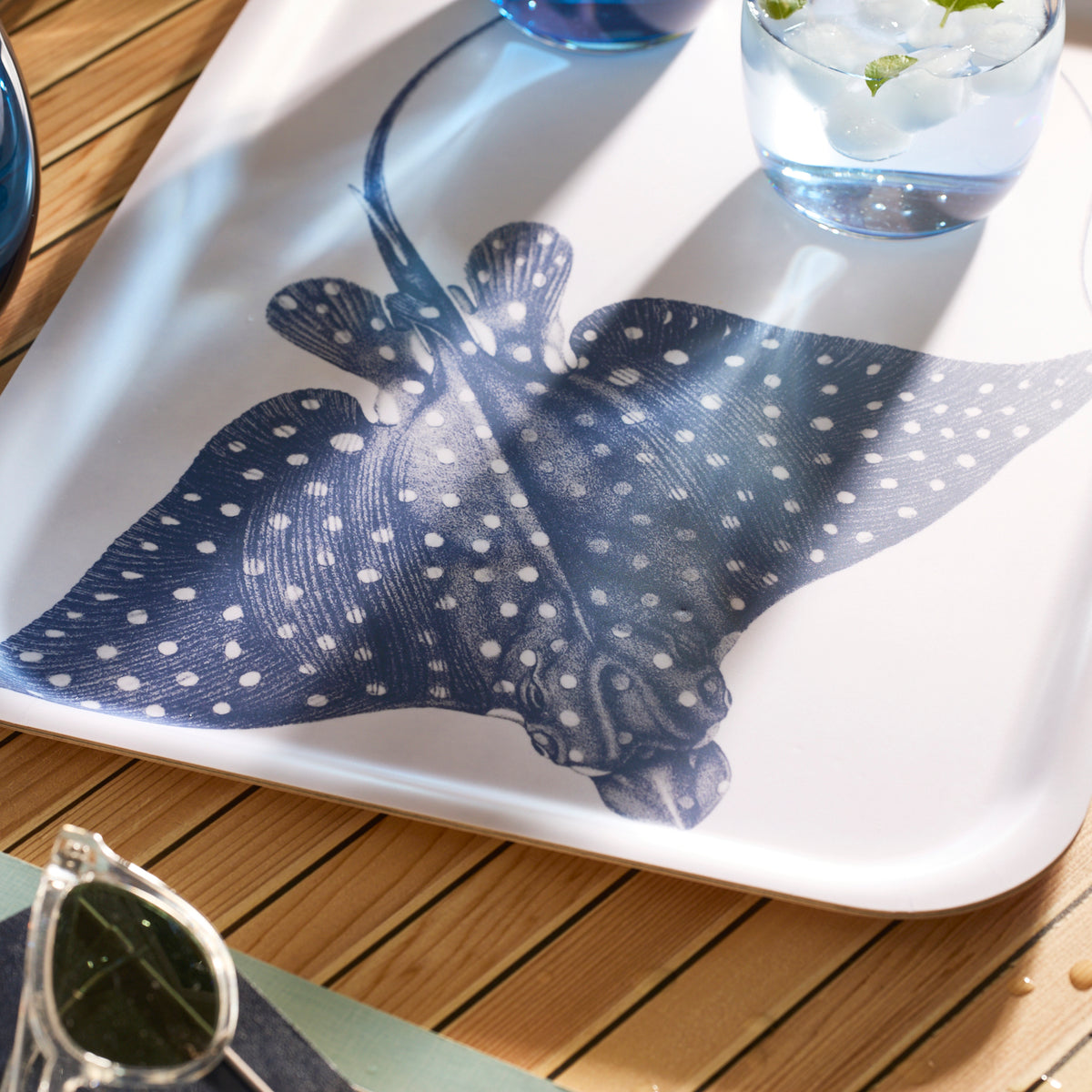 The Spotted Stingray Large Rectangular Birchwood Tray from Caskata&#39;s Coastal Collection, showcasing a blue and white stingray design, sits on a wooden surface alongside a refreshing glass of ice water and trendy sunglasses.