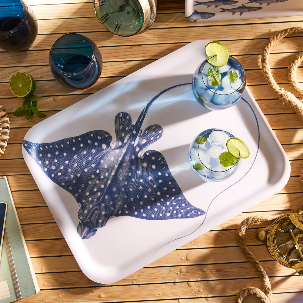 The Spotted Stingray Large Rectangular Birchwood Tray by Caskata elegantly holds two glasses with a clear beverage and lime slices. Positioned on a wooden table with a rope, lime slice, and open book, it adds charm to any setting.