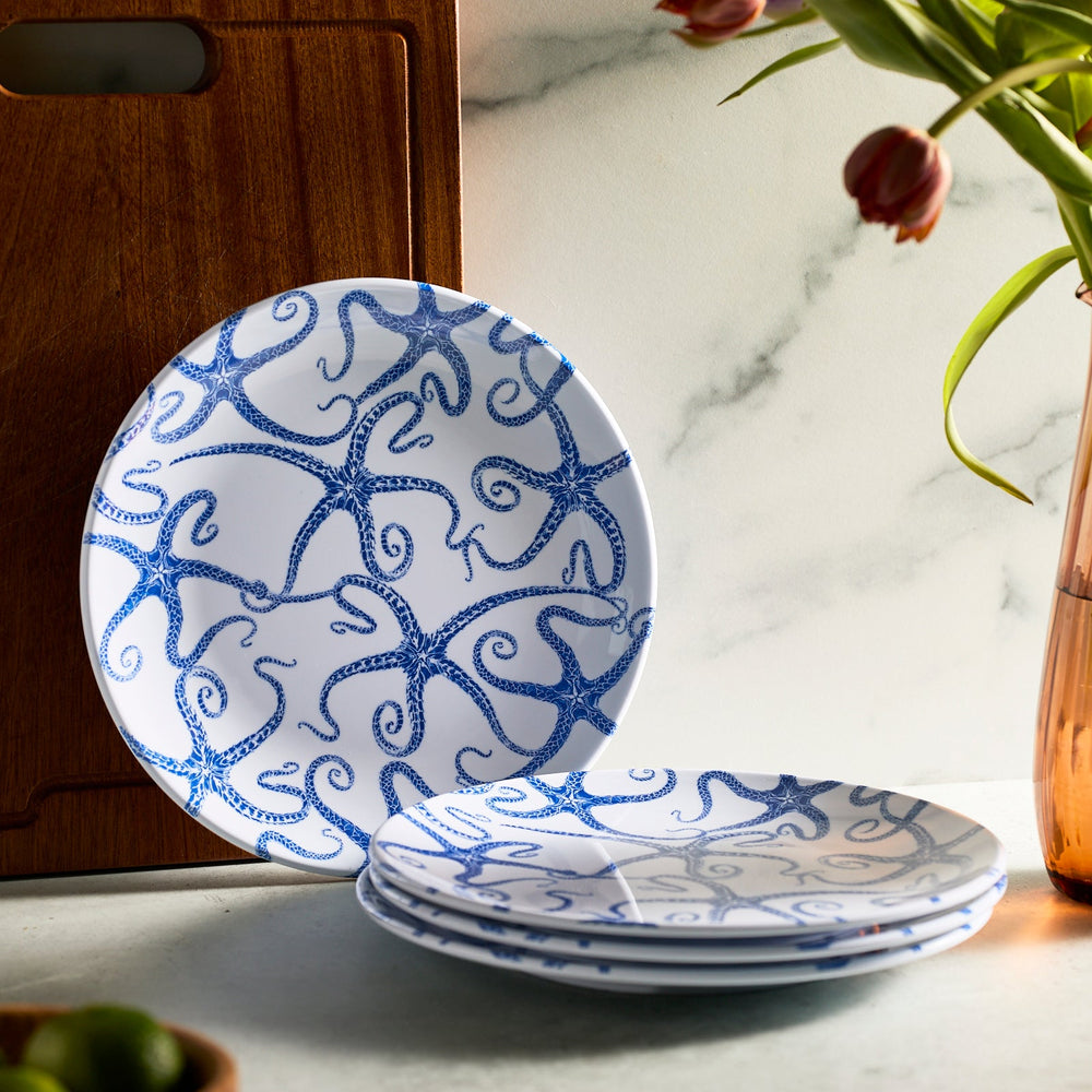 Caskata's Starfish Melamine Dinner Plates, Set of 4, featuring a blue starfish pattern, are stacked beside a wooden board. Tulips in a vase and limes accent the marble-like surface. Crafted from BPA-free melamine, these plates blend style and safety for your dining experience.