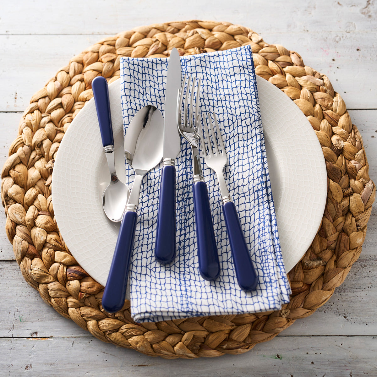 A place setting with a white plate, a Caskata Catch Dinner Napkins, Set of 4 100% cotton napkin, blue-handled utensils, and a woven placemat on a white wooden surface.