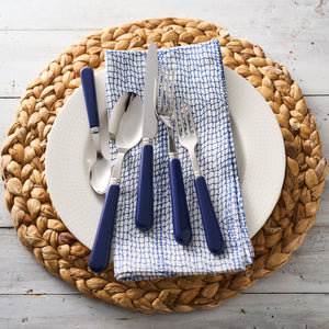 A place setting with a white plate, a Caskata Catch Dinner Napkins, Set of 4 100% cotton napkin, blue-handled utensils, and a woven placemat on a white wooden surface.