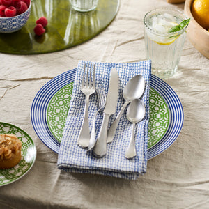 A place setting with a fork, knife, and two spoons rests on a blue-patterned, 100% cotton Catch Dinner Napkins, Set of 4 by Caskata atop a green and white plate. Nearby sits a drink with ice and a lemon slice, accompanied by a small dish of raspberries.