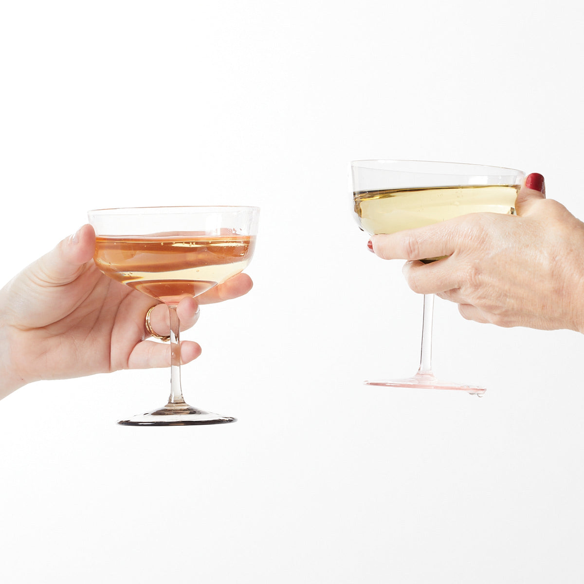 A pair of Celia Rose &amp; Mocha Coupe Cocktail Glasses by Caskata filled with wine, held by hands.