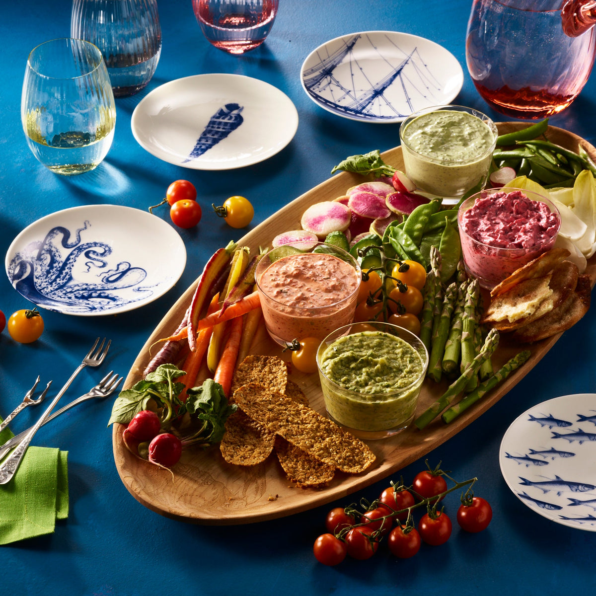 A wooden platter with colorful vegetable dips and fresh vegetables is surrounded by small, illustrated Caskata Artisanal Home Lucy Small Plates, glassware, and utensils on a blue surface.
