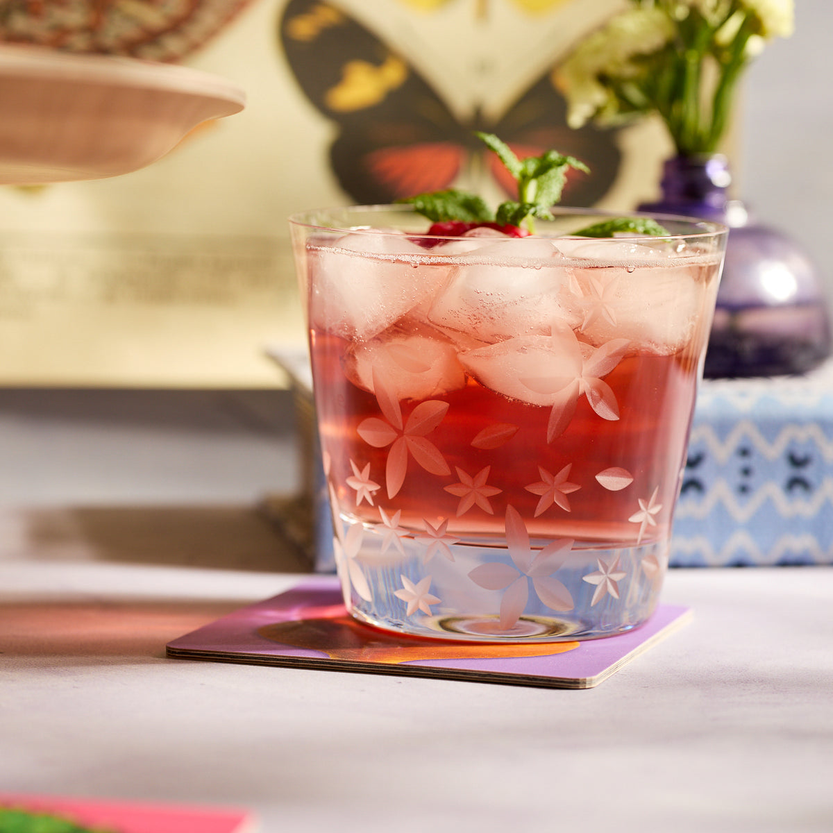 A glass of pink iced drink with mint, served on a colorful coaster, rests on a floral and butterfly-themed table, crafted from Caskata’s Chatham Bloom Tumblers—lead-free crystal inspired by the Collection’s elegant floral pattern.