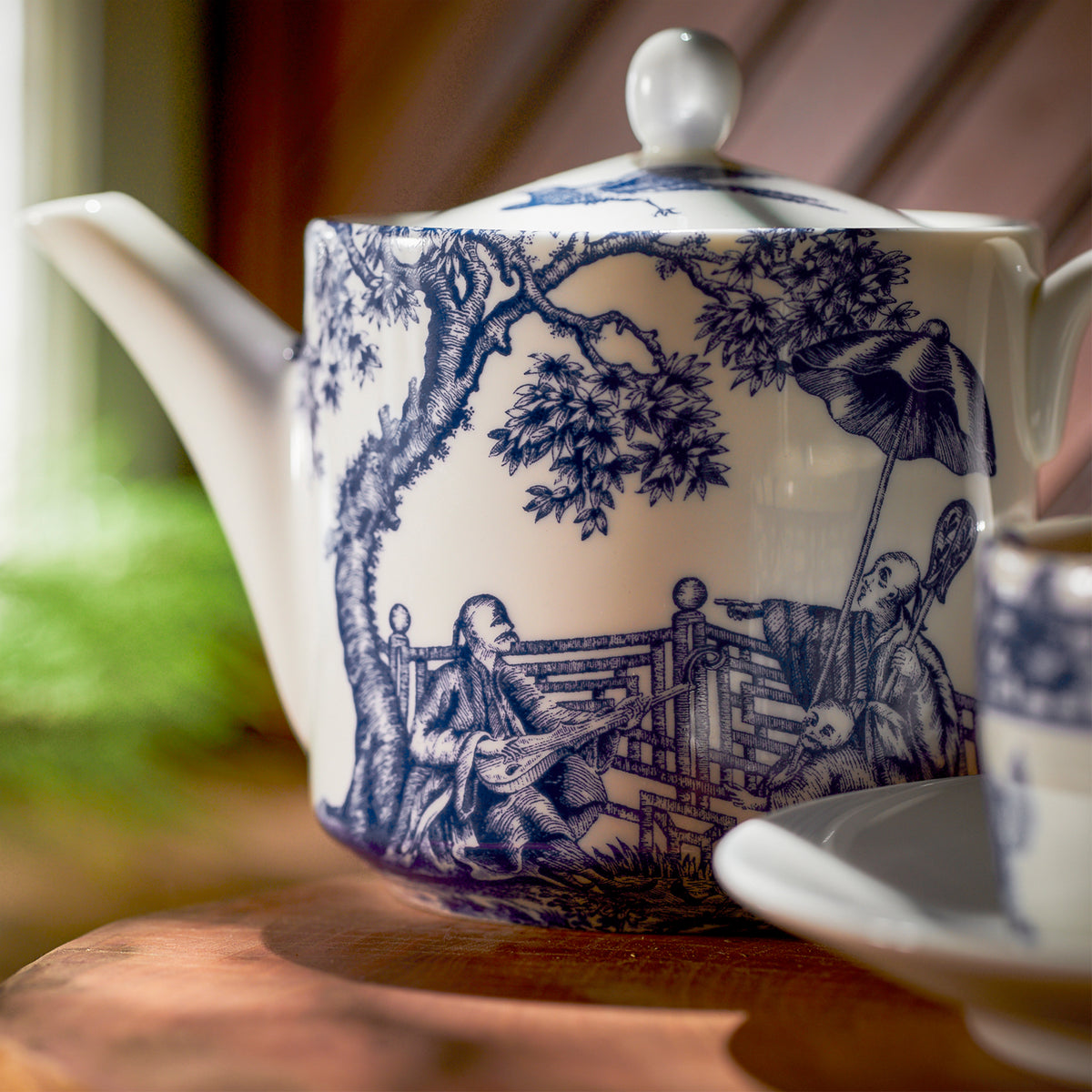 The Caskata Chinoiserie Tea Bundle showcases a porcelain teapot adorned with blue pastoral scenes, including two people, a tree, and a bridge. It sits elegantly on wood next to a matching teacup.