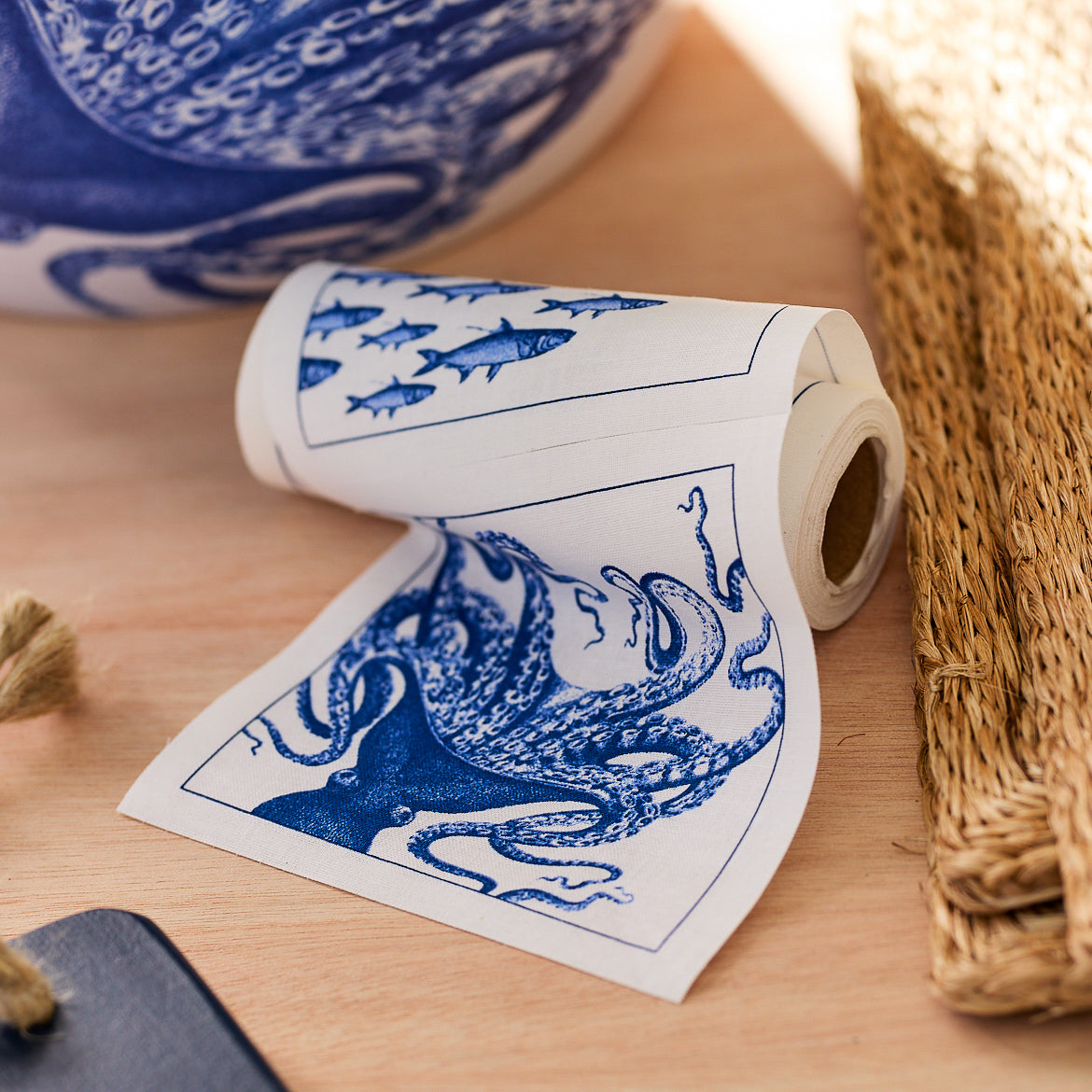 A roll of Coastal Cocktail Napkin Roll by MY DRAP with blue coastal patterns featuring illustrations of an octopus and fish, partially unrolled on a wooden surface next to a woven mat.