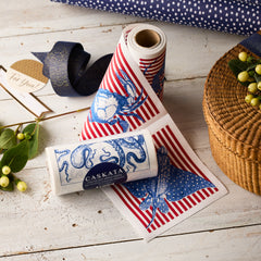 The MY DRAP Mixed Coastal Cocktail Napkin Rolls, Set of 2, feature blue sea creatures and red stripes—perfect for adding a coastal touch to your next gathering. Displayed with a wicker basket, ribbon, greenery, and a gift tag on a white wooden table.